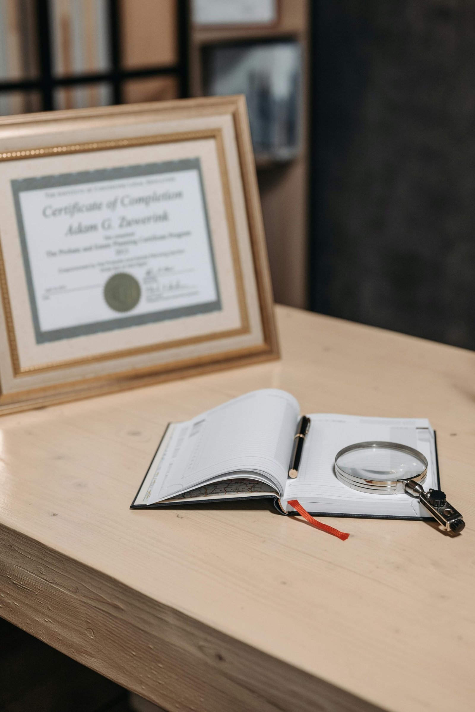 Open notebook with magnifying glass on wooden table beside framed certificate.
